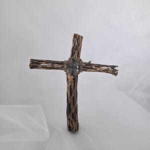 May include: A rustic wooden cross with a natural, textured appearance. The cross is constructed from dried branches, with a central knot detail. The color is a mix of brown and tan, with a light-colored background.