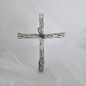 May include: A handcrafted cross made from a textured, light-colored material, possibly wood, with dark accents. A small, purple gemstone is set at the intersection of the cross. The cross is displayed against a white background.