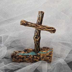 May include: A cross sculpture crafted from textured brown cholla wood, featuring a horizontal crossbar attached to a vertical post. The base is a thick, cylindrical piece of wood with a central groove filled with small, bright turquoise stones.
