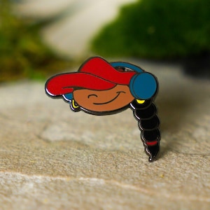May include: Enamel pin featuring a smiling cartoon character with brown skin, wearing a red hat, blue headphones, and a black braid. The pin has a black outline and gold earrings. The pin is on a light brown surface.