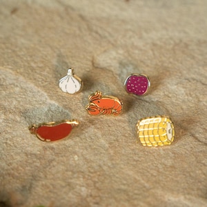 May include: A collection of enamel pins featuring food items. The pins include a garlic clove, a lobster, a raspberry, a sausage, and a sushi roll. Each pin has gold-colored trim and is set against a textured, neutral background.