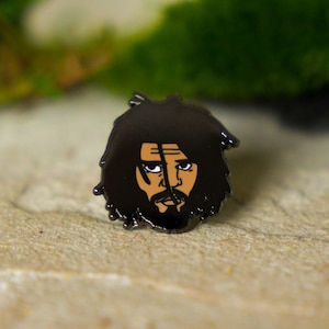 Könnte beinhalten: Schwarzer Emaille-Pin mit einem stilisierten Gesicht mit dunklem Haar und Bart. Das Gesicht hat gebräunte Haut, einen Schnurrbart und einen Spitzbart. Der Pin ist vor einem neutralen Hintergrund platziert, ideal für Kleidung.