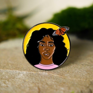 May include: A round enamel pin featuring a woman with dark skin and black curly hair, set against a yellow background. A monarch butterfly rests on her hair. The pin has a black border and a pink shirt.