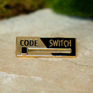 May include: A rectangular gold-coloured pin with the words "CODE SWITCH" in black lettering. The pin has a sliding bar design, with a black section on the right and a gold section on the left. It's likely a lapel pin or accessory.