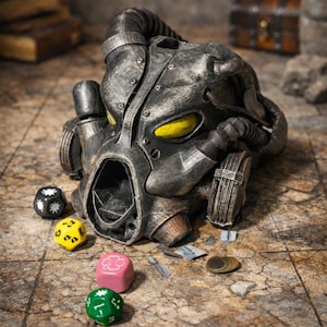 Puede incluir: Un casco gris metálico detallado con lentes oculares amarillas, que recuerda una estética postapocalíptica. Varios dados coloridos, incluyendo negro, amarillo, rosa y verde, están dispersos cerca. También hay una moneda y pequeños trozos de metal.