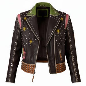 Puede incluir: Chaqueta de cuero marrón con cuello verde y rayas rosas en los hombros. La chaqueta está adornada con tachuelas plateadas y diseños de estrellas amarillas. El dobladillo y los puños tienen detalles de cuero marrón con tachuelas. Cierre de cremallera y tirador de cuero.