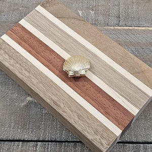 May include: A rectangular wooden box featuring alternating horizontal stripes of light brown, dark brown, and cream wood grain. The lid is adorned with a centered, metallic gold-toned seashell-shaped knob, providing a decorative accent to the surface.