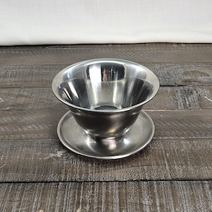 May include: A polished stainless steel bowl with a wide, flared rim resting on a matching circular saucer. The metal surface is reflective and smooth, featuring a deep, rounded basin that tapers toward the base, creating a clean, functional design.