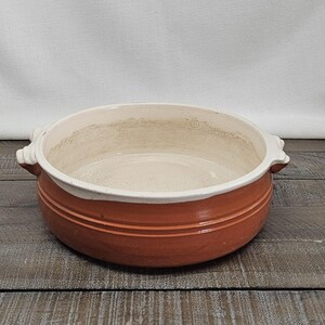 May include: A round, terracotta-colored ceramic bowl with a white interior and two small handles. The bowl has a wide rim and two horizontal grooves around the exterior. The bowl is sitting on a wooden surface.