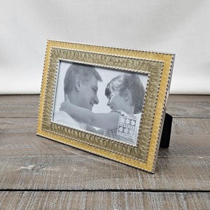 May include: A rectangular picture frame with a textured gold-toned border and a beaded inner edge. The frame contains a black and white photograph of a person embracing a child, with the text SIXTREES visible in the bottom right corner of the photo.