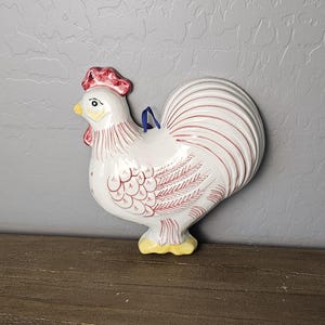 May include: A white ceramic wall plaque shaped like a rooster, featuring a red comb and wattle, yellow beak and feet, and fine red line detailing on the feathers. A small blue ribbon loop is attached to the back for hanging purposes.