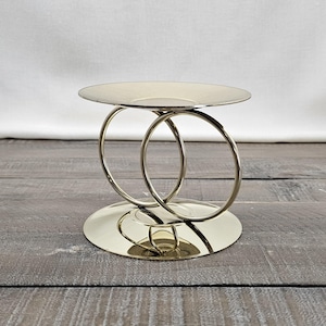 May include: A gold-toned metal candle holder featuring a flat circular base and a matching circular top plate. The two components are connected by two interlocking vertical rings that create a structural support for the top platform.