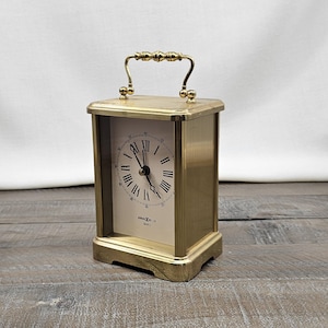 May include: A rectangular brass-colored clock with a handle. The clock face has black Roman numerals and hands. The clock is sitting on a wooden surface.