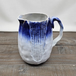 May include: A ceramic pitcher with a corn-on-the-cob design. The pitcher features a white base with a blue ombre effect on the top and handle. The corn design is textured, adding visual interest. The pitcher is set against a wooden surface.