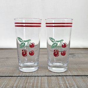 May include: Two clear drinking glasses with a red cherry design. Each glass has two red stripes near the top and a cluster of red cherries with green leaves. The glasses are on a wooden surface.