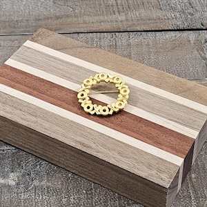 May include: A circular gold-toned brooch featuring a repeating XOXO pattern, resting on a rectangular wooden box constructed from alternating light, medium, and dark brown wood strips with a visible grain and smooth finish.