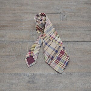 May include: A necktie featuring a multi-colored plaid pattern with intersecting lines in shades of beige, burgundy, blue, and yellow. The fabric has a visible woven texture, and the tie is folded to display the front pattern and the solid burgundy lining.