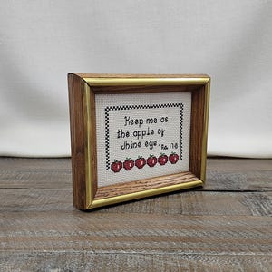 May include: A framed cross-stitch piece featuring the black text "Keep me as the apple of thine eye Ps. 17:8" surrounded by a black checkered border and a row of six red apples with green stems, encased in a wooden frame with gold-toned trim.