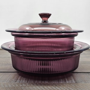 May include: A set of two round, deep purple glass baking dishes with vertical ribbed sides. The top dish features a matching glass lid with a central knob handle. Both dishes have wide, flat rims and a translucent, deep violet color throughout.