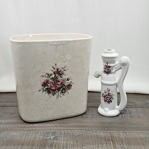 May include: A cream-colored ceramic wastebasket and a water pump-shaped vase. Both items feature a floral design of burgundy roses. The wastebasket has a speckled pattern, while the vase has a handle and spout.