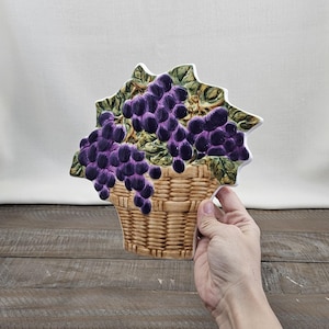 May include: A ceramic wall plaque shaped like a woven wicker basket overflowing with clusters of deep purple grapes and green leaves. The piece features a textured, three-dimensional design with a glossy finish, showcasing detailed grape bunches.