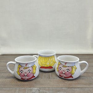 May include: Three white ceramic mugs with rounded bodies and handles. Two mugs feature a cartoon face with blonde hair and a white cap with a red letter C. The center mug shows the back of the blonde hair with a red collar detail on the base.
