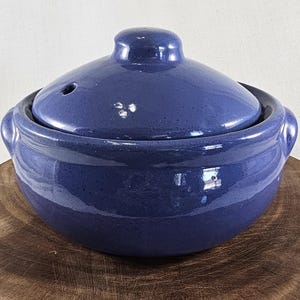 May include: A round, deep blue ceramic casserole dish with a matching lid. The pot features a glossy, speckled finish, two integrated side handles, and a small steam vent hole located on the top of the domed lid. The surface is smooth and reflective.