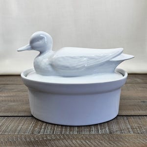 May include: A white ceramic duck-shaped dish with a lid. The duck figurine sits atop the lid, with detailed feather and wing designs. The dish is a light blue color and sits on a wooden surface.