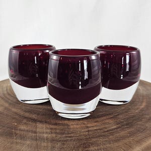 May include: Three stemless wine glasses with a deep burgundy color and a clear, white base. The glasses are cylindrical with a rounded bottom and are arranged on a wooden surface. The glasses are likely for serving wine or other beverages.
