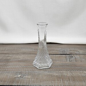 May include: A clear glass bud vase with a textured, bubble-like pattern on the lower portion and a smooth rim. The vase sits on a weathered wooden surface, with a white fabric backdrop.