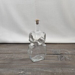 May include: A clear glass bottle shaped like a violin or cello, featuring a slender neck and a rounded body with curved sides. The bottle is topped with a small, cylindrical cork stopper that fits securely into the narrow opening at the top.