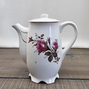 May include: White ceramic teapot with a lid and handle. The teapot features a floral design with pink roses, green leaves, and brown stems. The spout has a gold trim. The teapot sits on a wooden surface.