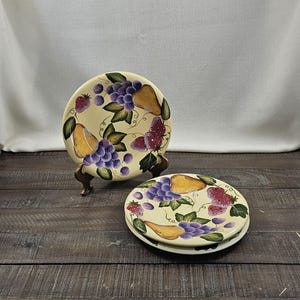 May include: Three round ceramic plates featuring a hand-painted fruit motif. The design includes clusters of purple grapes, yellow pears, and red strawberries with green leaves on a cream-colored background. One plate rests on a metal stand.