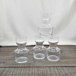 May include: A collection of clear glass carafes, some stacked, on a wooden surface. The carafes have a unique hourglass shape with a wide base and a narrow neck. One carafe has the text "0.25 l SAIVO" printed on it.