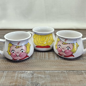 May include: Three white ceramic mugs with handles, each featuring a cartoon image of a blonde girl with rosy cheeks, a red scarf, and a white cap. The mugs are on a wooden surface.