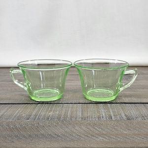 May include: Two clear, green-tinted glass teacups with handles. The cups have a rounded shape and sit on a wooden surface. The glass has a slight glow, suggesting a vintage or antique style. The cups are empty.