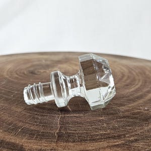 May include: A clear, faceted glass bottle stopper with a threaded base and a geometric, diamond-shaped top. The stopper is lying on a dark brown, wooden surface.