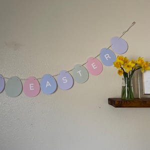 May include: A pastel Easter banner spelling "EASTER" with egg-shaped cutouts strung on a thin string. A small glass vase with yellow daffodils sits on a wooden shelf, adding a touch of spring.