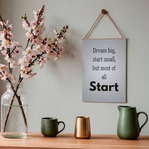 May include: A still life arrangement featuring a clear glass vase with pink flowering branches, a green mug, a gold-colored cylindrical container, and a green pitcher. A framed sign with the text "Dream big, start small, but most of all Start" hangs on the wall.
