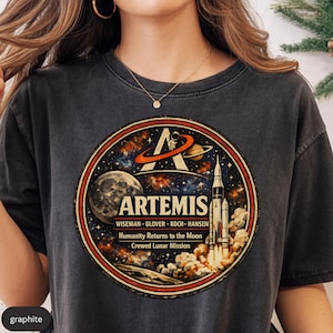 May include: Graphite-colored t-shirt featuring a circular graphic with the word "ARTEMIS" and a space-themed design. The graphic includes a rocket launching, a moon, and the text "WISEMAN - GLOVER - KOCH - HANSEN Humanity Returns to the Moon Crewed Lunar Mission."