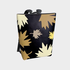 May include: A black backpack with a repeating pattern of stylized leaves in shades of cream, tan, and gold. The backpack has black straps and a top handle. The leaves are arranged in a random pattern across the surface of the bag.