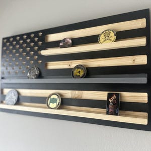 May include: A wooden American flag-shaped display shelf with black and natural wood tones. The shelf has multiple horizontal slots to hold challenge coins. Several coins are displayed on the shelves, showcasing various designs and colors.