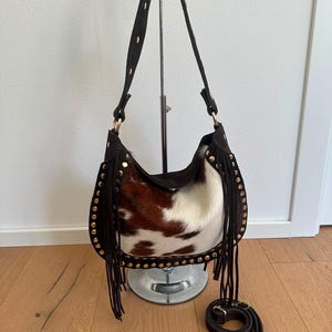 May include: A brown and white cowhide shoulder bag featuring long dark brown fringe along the sides and bottom. The bag is accented with gold-tone metal studs along the edges and includes a matching adjustable shoulder strap with gold hardware.