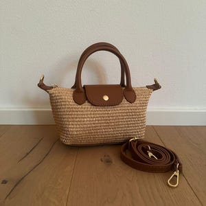 Pode incluir: Uma pequena mala com corpo em palha entrançada em tom bege natural, acentuada com pegas em pele castanha e uma aba a condizer com um botão de pressão dourado. Uma alça de ombro em pele castanha destacável está enrolada ao lado da mala.