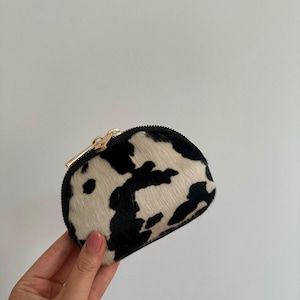 Peut inclure: Un petit porte-monnaie semi-circulaire avec un motif de vache noir et blanc et une finition texturée en poils. La pochette est fermée par une fermeture éclair noire dotée d'une tirette en métal doré brillant.