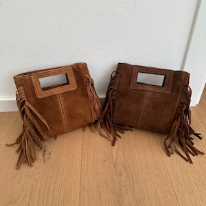May include: Two suede handbags with rectangular cut-out handles and long fringe detailing along the sides. One bag is a light tan color, while the other is a darker chocolate brown. Both feature a vertical seam down the center of the front panel.
