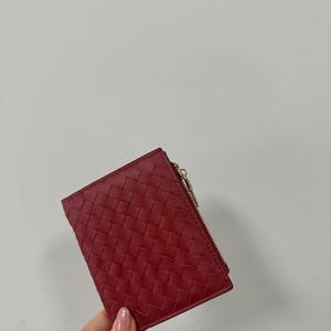 Może przedstawiać: Mały, czerwony portfel z plecionym wzorem. Portfel ma złote zapięcie na zamek błyskawiczny. Projekt jest klasyczny i minimalistyczny. Portfel jest trzymany w dłoni osoby.
