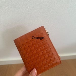 Może przedstawiać: Pomarańczowy sk&oacute;rzany portfel z tkanym wzorem. Portfel ma zapięcie na zamek błyskawiczny, a na przodzie nadrukowano napis "Orange". Portfel jest trzymany w dłoni.