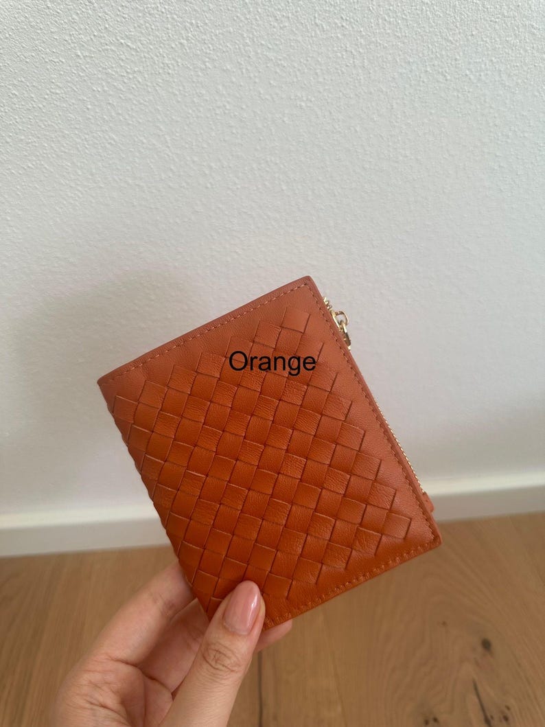 Może przedstawiać: Pomarańczowy sk&oacute;rzany portfel z tkanym wzorem. Portfel ma zapięcie na zamek błyskawiczny, a na przodzie nadrukowano napis "Orange". Portfel jest trzymany w dłoni.