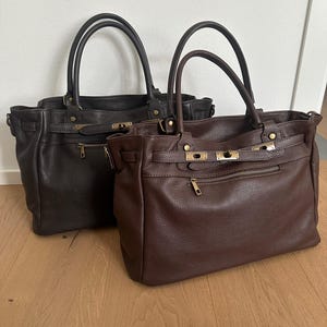 May include: Two structured handbags made of textured leather, one in dark brown and one in black. Each bag features dual top handles, a horizontal front zip pocket, and a decorative strap closure with metallic hardware accents across the front.